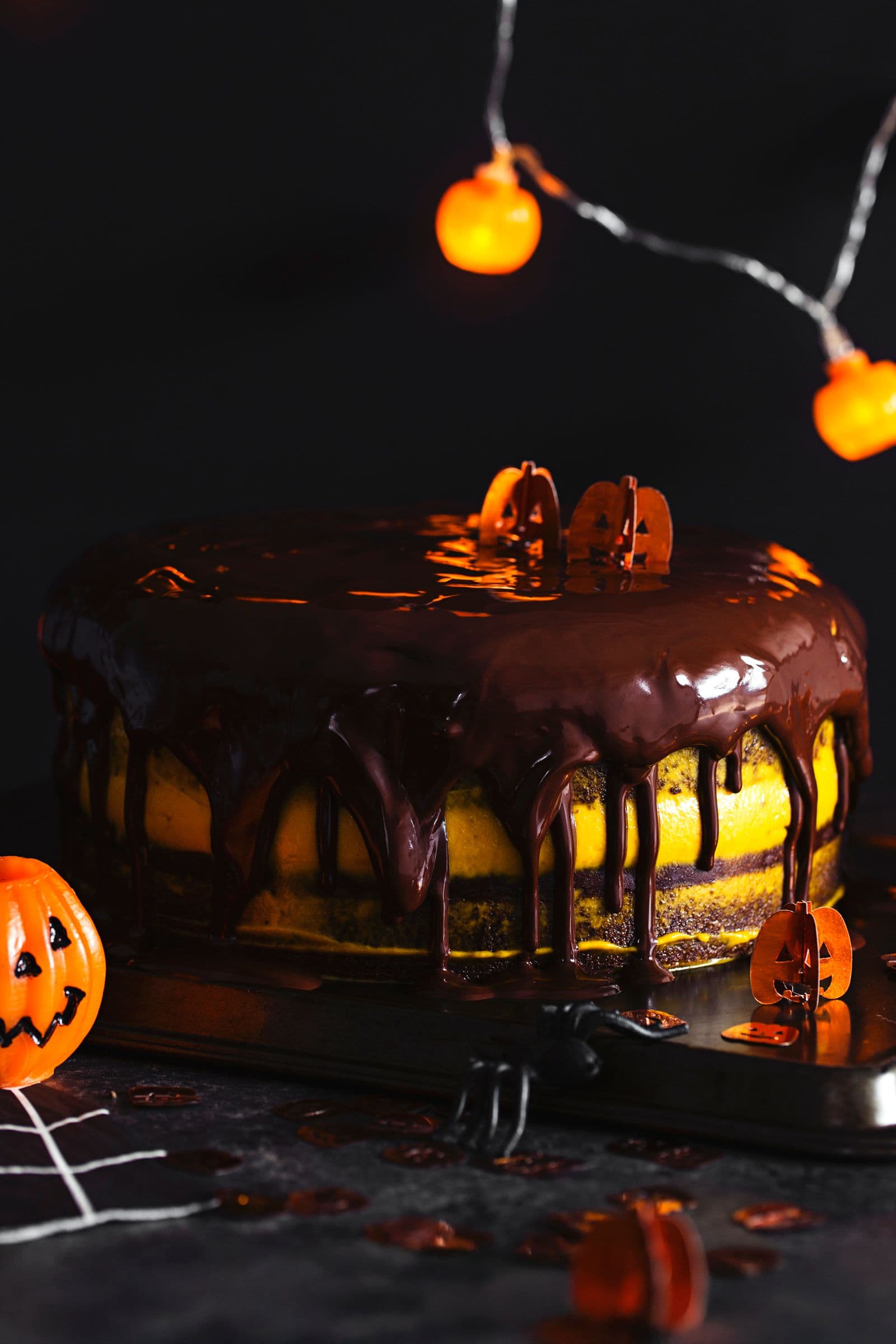 Halloween layered cake with dripping chocolate glaze, topped with tiny pumpkin decorations and orange lights in the background.