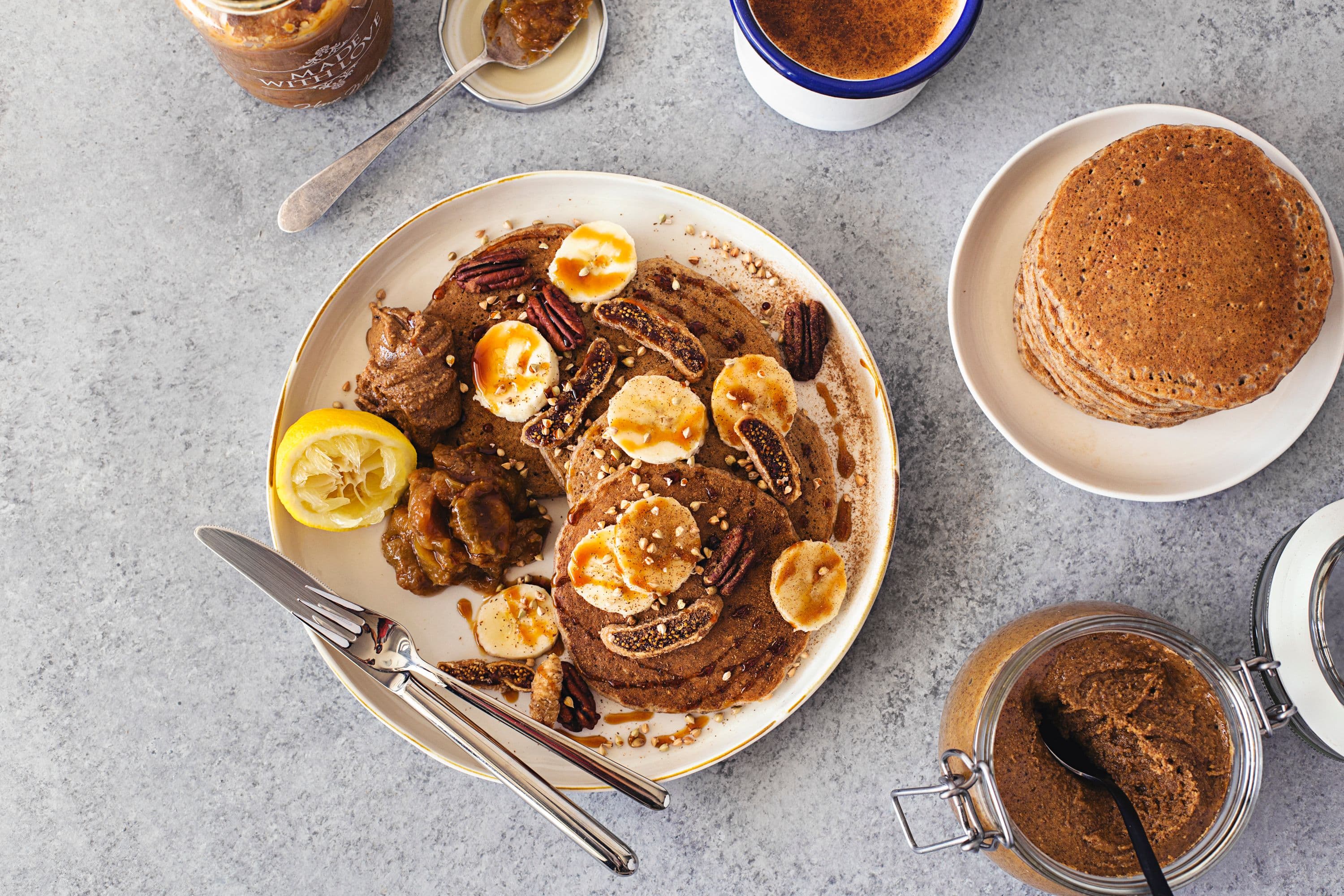 Tabletop flatlay with stacked pancakes, nut butter, jam, and sliced banana.