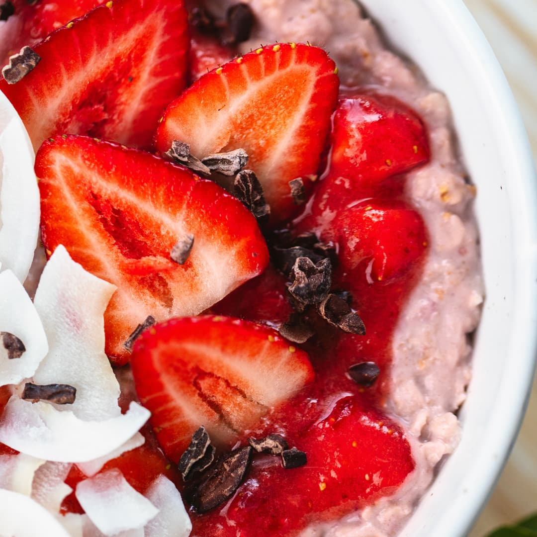 Pink, creamy oatmeal topped with sliced strawberries, strawberry sauce, coconut flakes and cacao nibs.