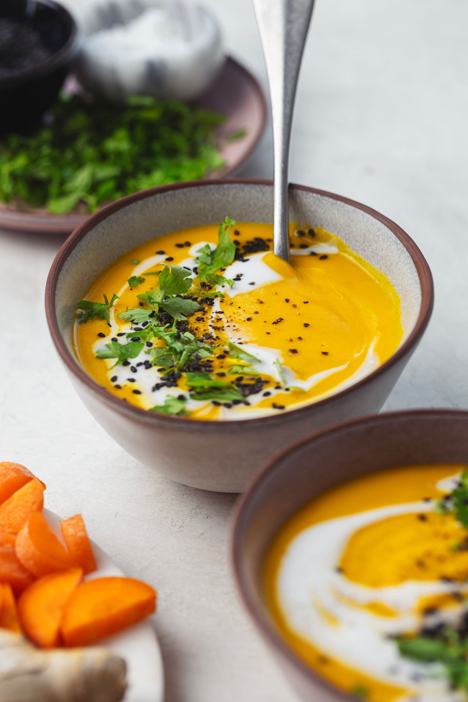 Taças de sopa de cenoura dourada com leite de coco em espiral, ervas frescas e sementes crocantes, prontas a servir.