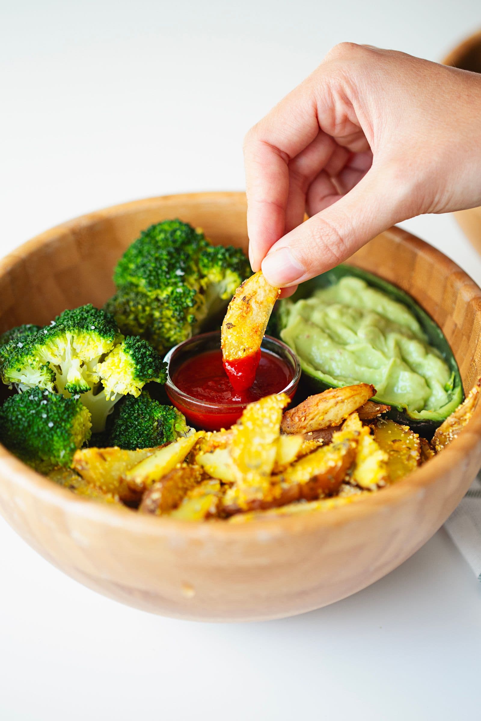 Mão a mergulhar gomo de batata crocante em ketchup numa taça com batatas e guacamole.