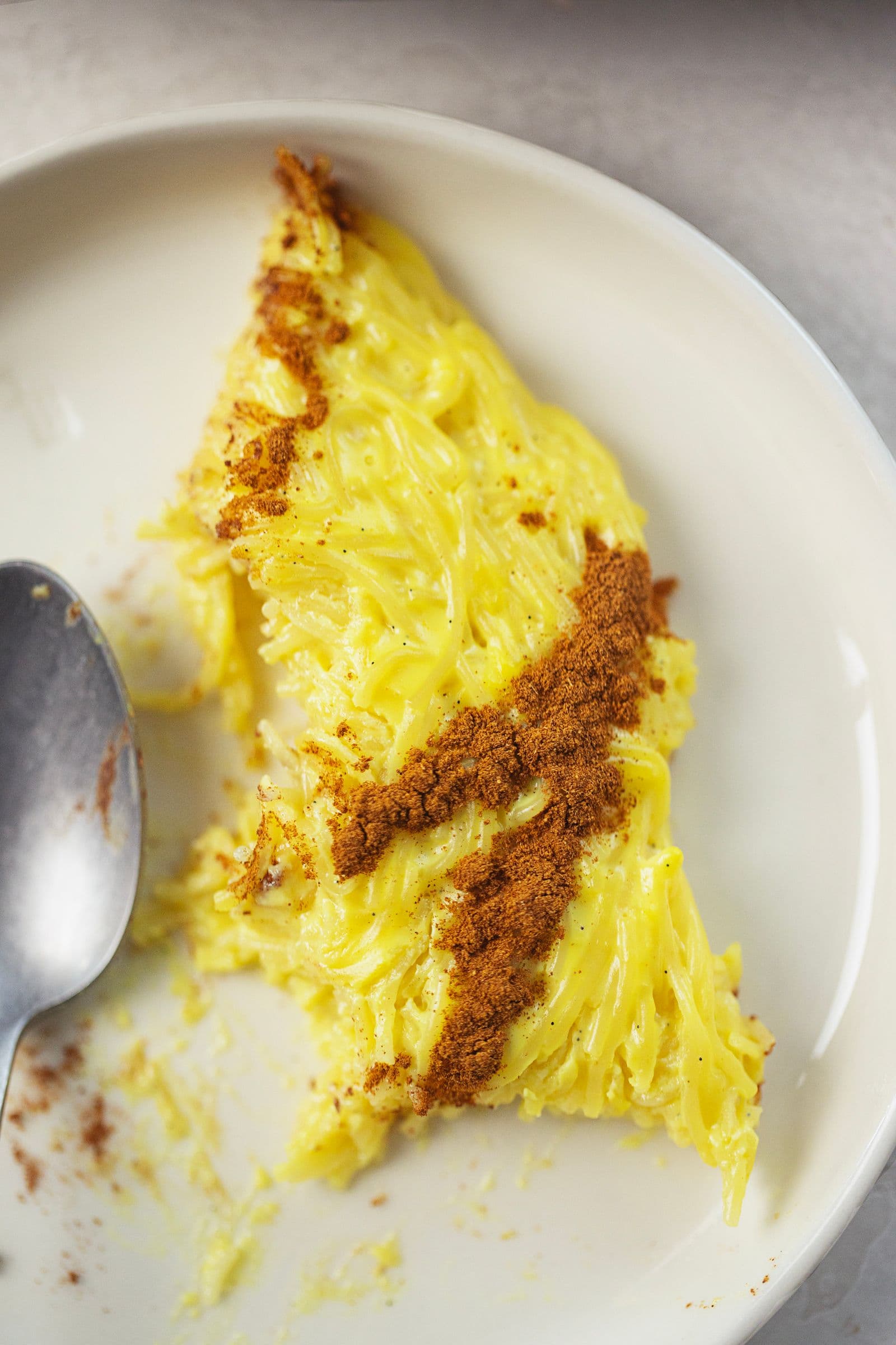 Fatia de aletria vegan parcialmente comida num prato branco com uma colher, a mostrar a textura cremosa e a cobertura de canela.