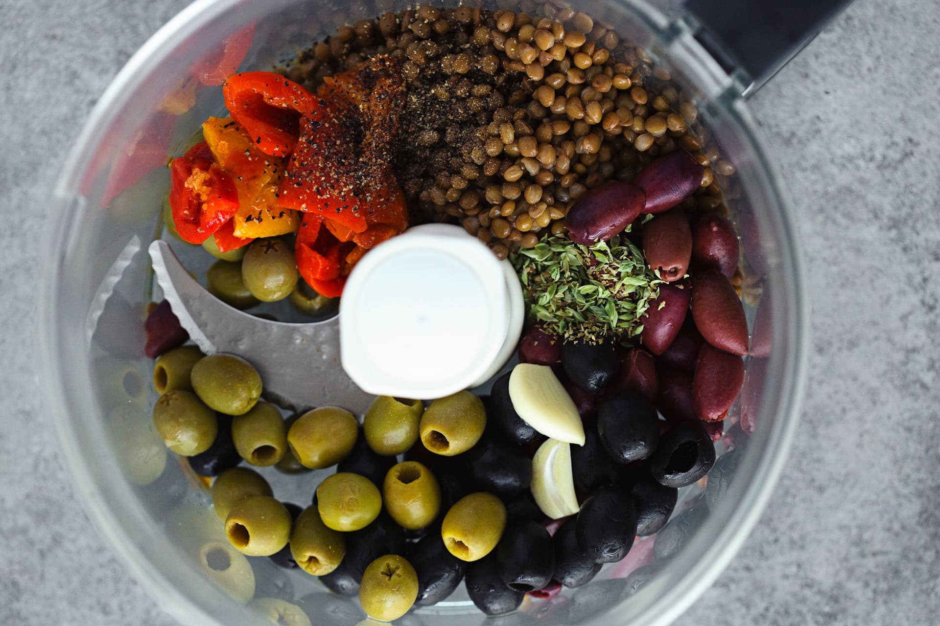 Bowl of a food processor filled with green and black olives, lentils, herbs and seasonings ready to make pâté.