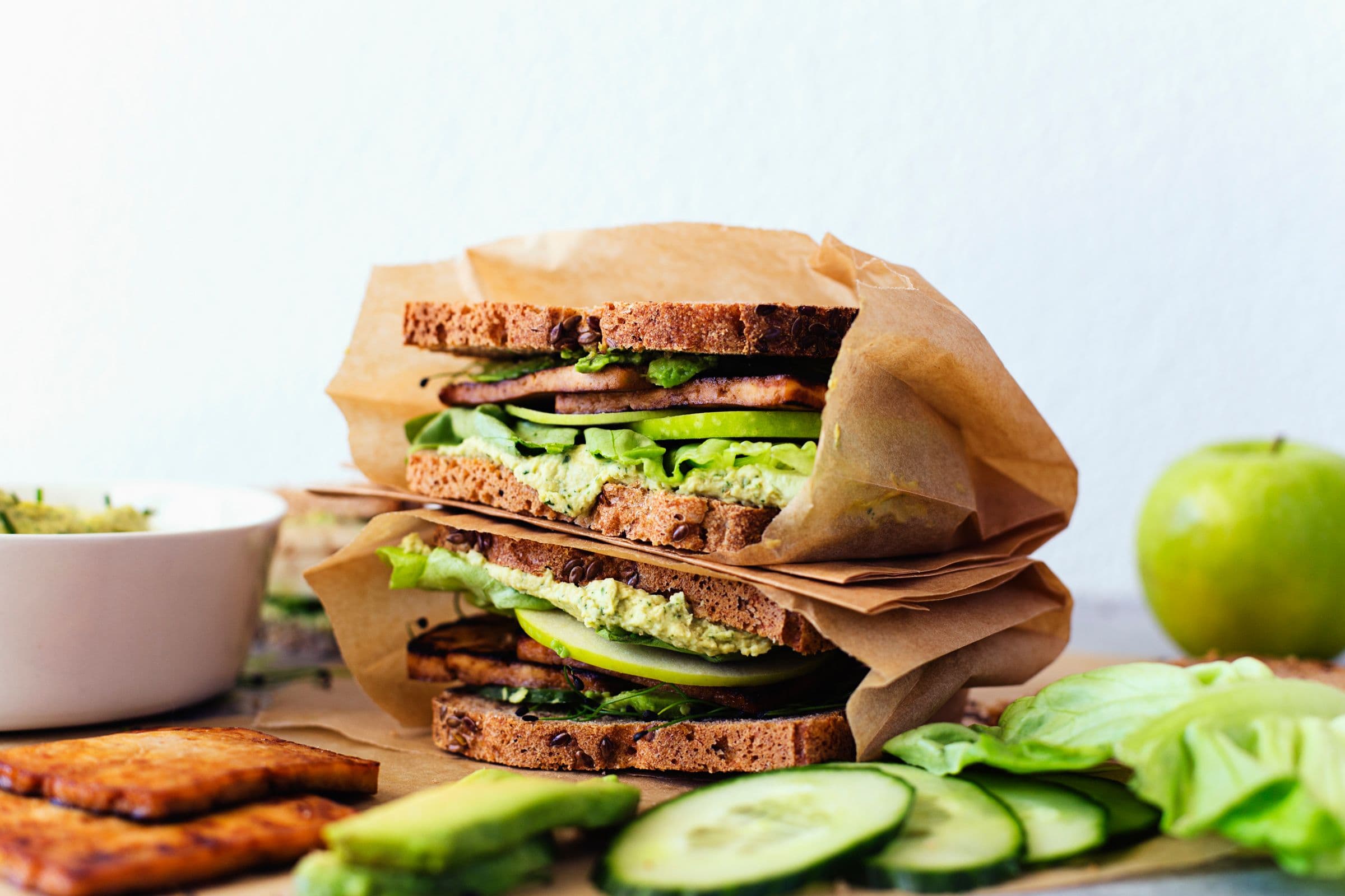 Vegan sandwiches with green vegetables, tofu, and herb hummus stacked in brown paper.
