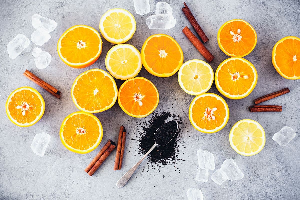 Slices of lemon and orange with cinnamon sticks, black tea leaves in and around a spoon and ice cubes.
