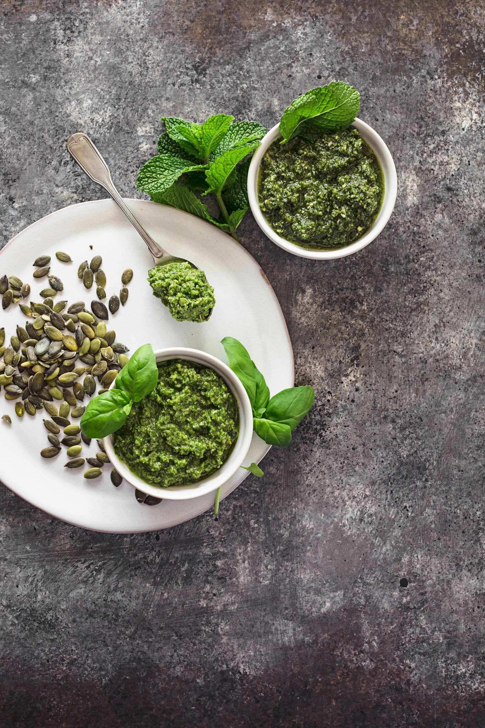 Two small bowls filled with homemade vegan pumpkin seed pesto, surrounded by fresh greens and seeds.