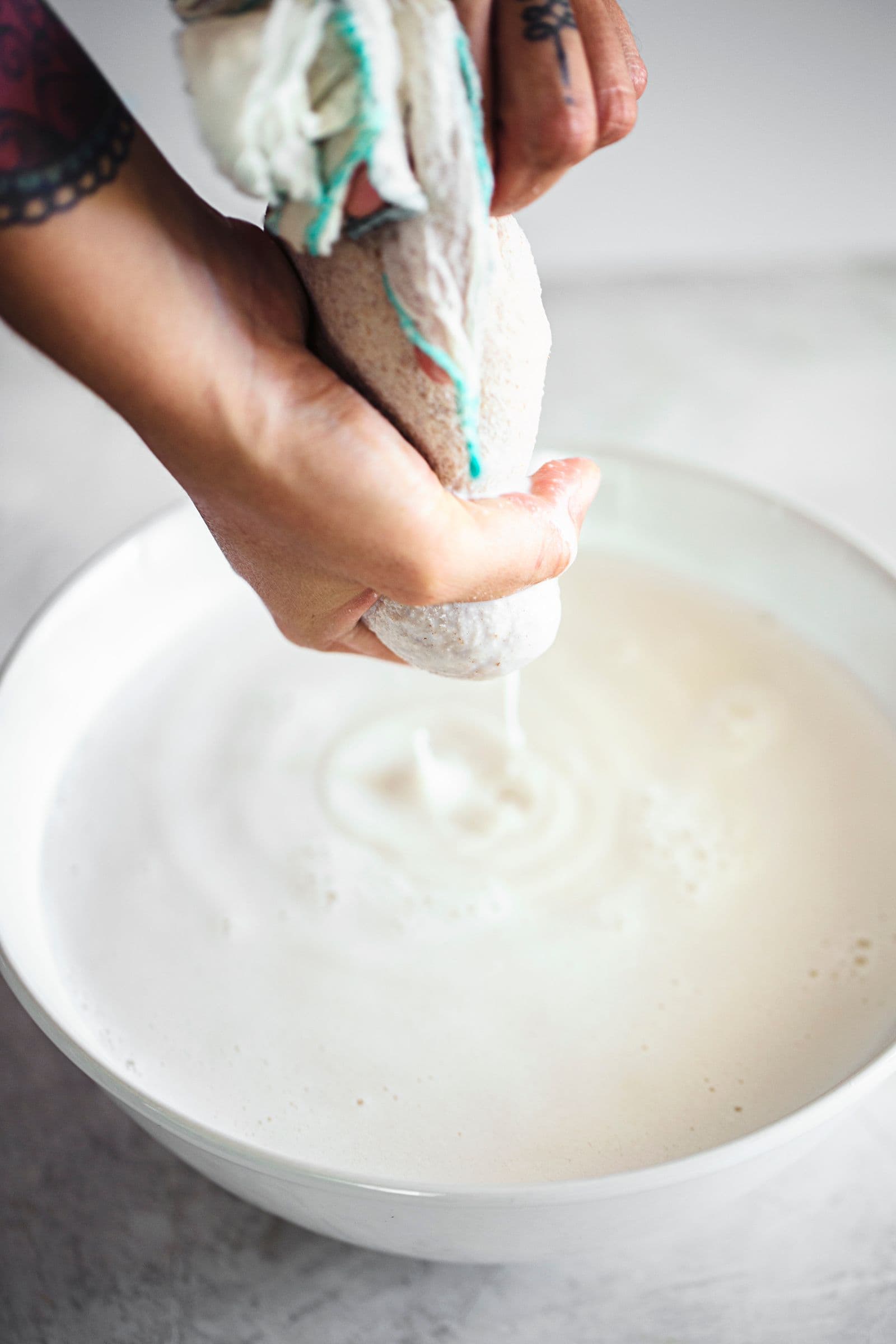 Hands squeezing homemade plant milk through a cheesecloth into a white bowl.