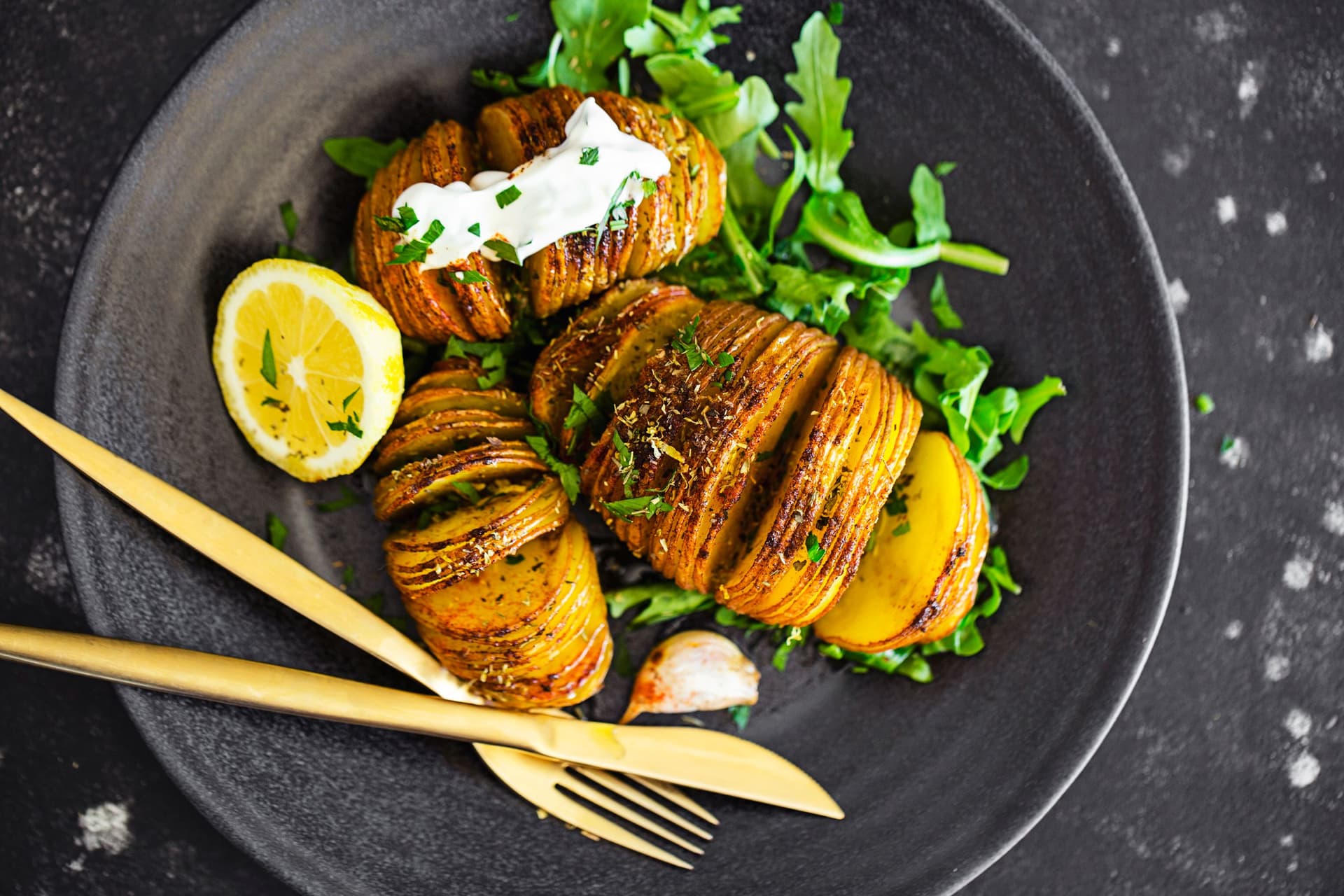Golden Hasselback potatoes with herbs, spices and creamy sauce, served with fresh arugula on a black plate.