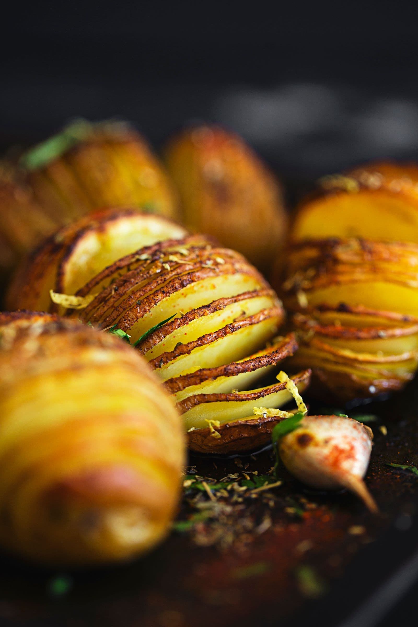 Close view of a seasoned Hasselback potato on a dark baking tray with roasted garlic.