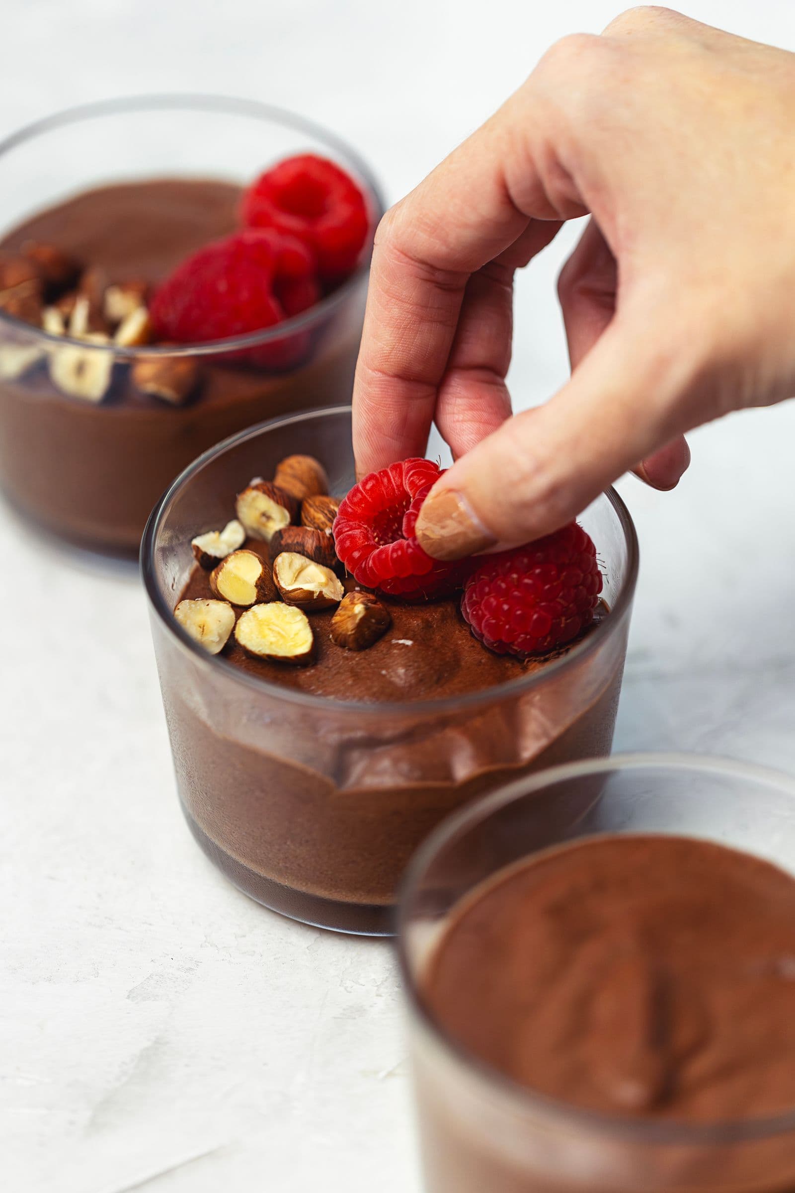 Mão a colocar framboesas frescas sobre a mousse de chocolate vegan num copo individual.