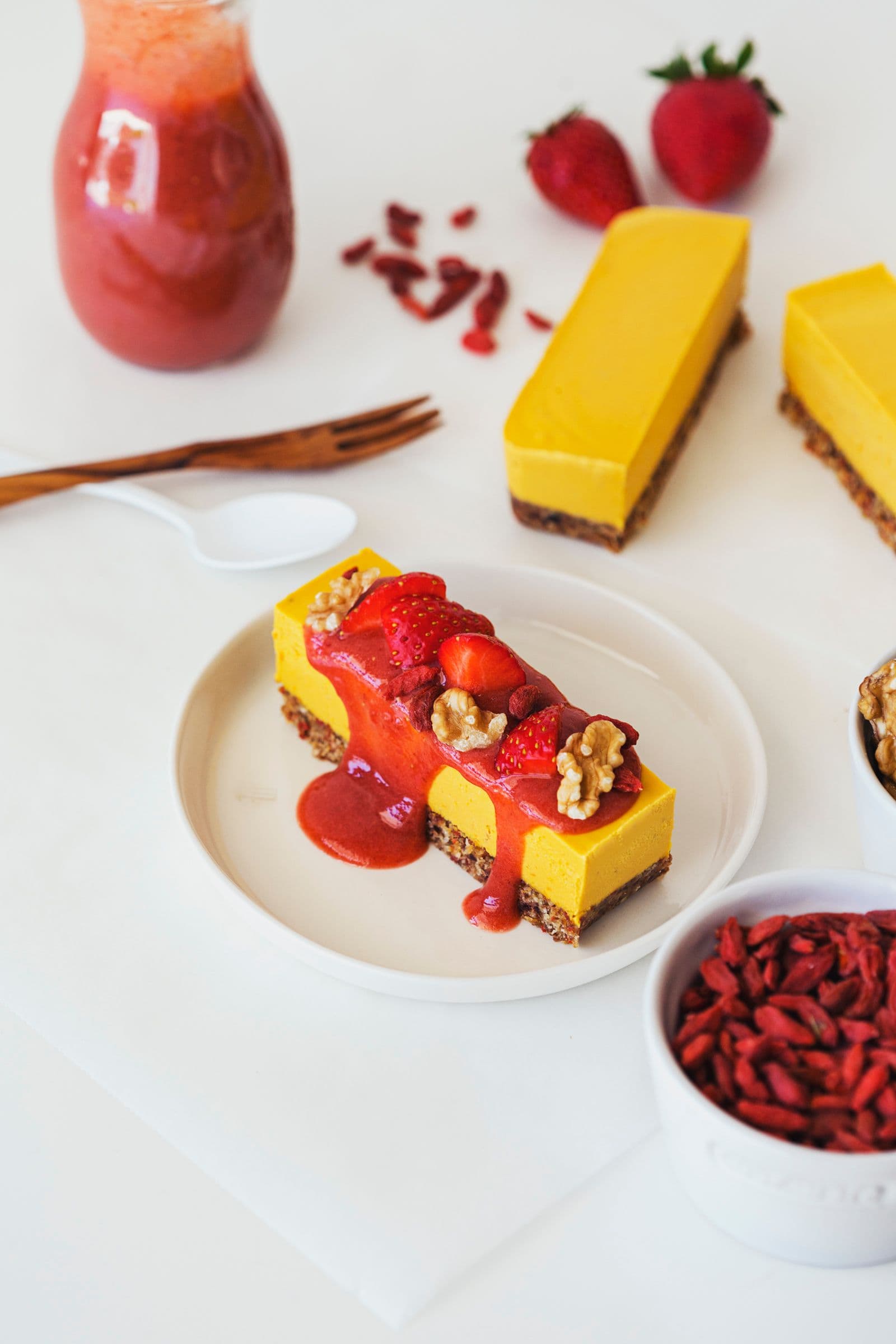 Uma fatia de cheesecake vegan de laranja e goji decorada com molho de morango, morangos frescos e nozes.