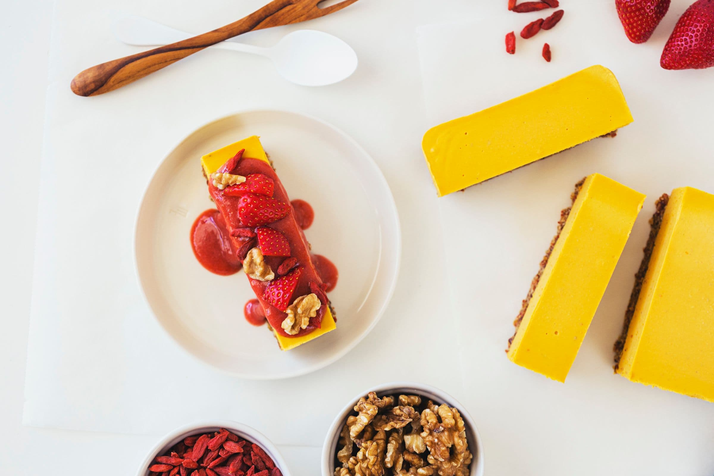 Slice of orange, goji, and strawberry raw vegan cheesecake topped with fresh strawberries, walnuts and sauce.