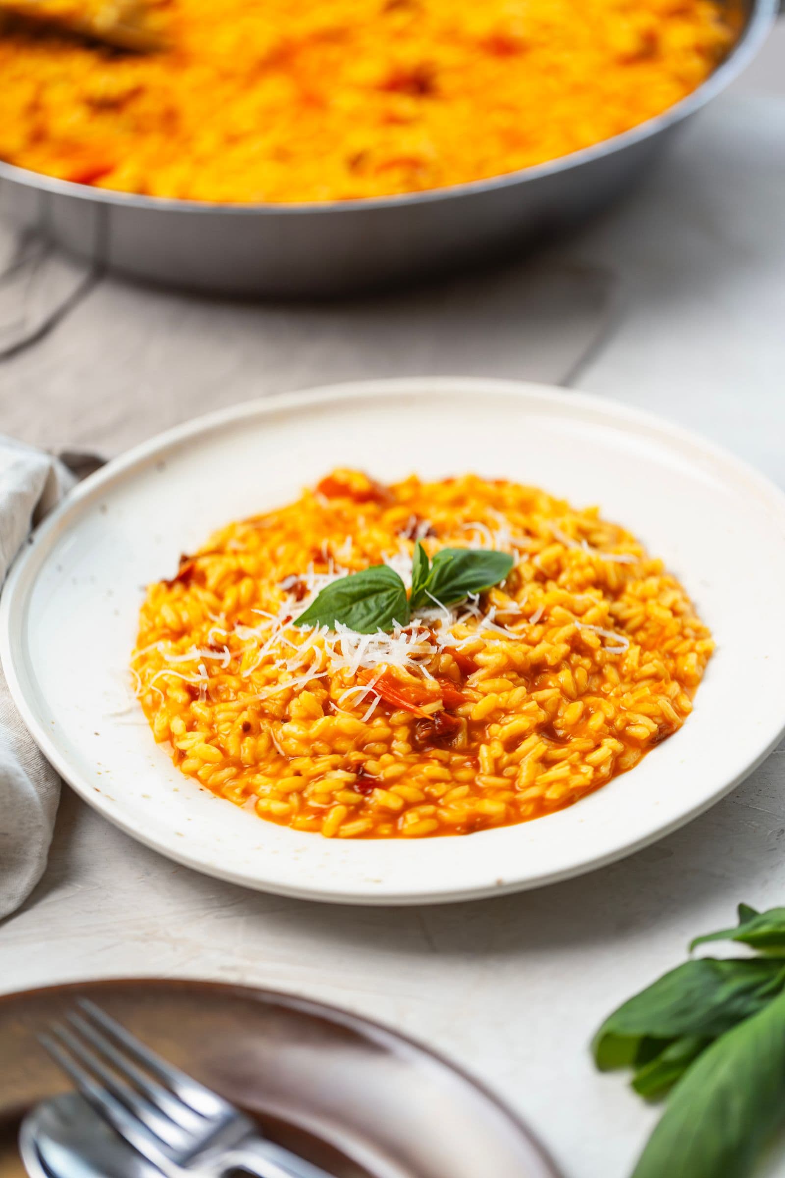 Risotto de tomate cremoso num prato branco, coberto com parmesão ralado e manjericão, com a frigideira ao fundo.