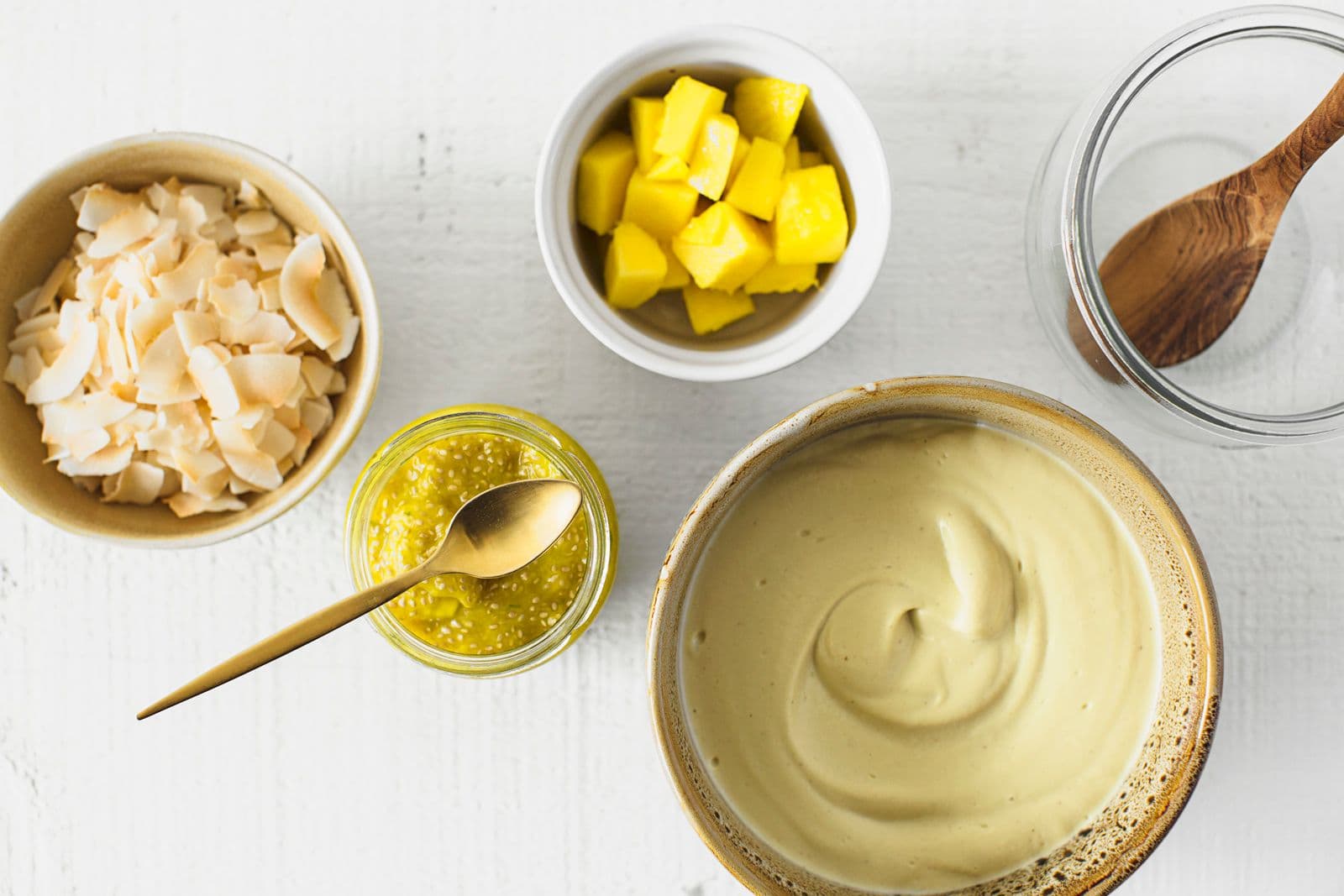 Taças pequenas com coco tostado, pedaços de manga fresca, doce de manga e chia e iogurte de caju e manga.